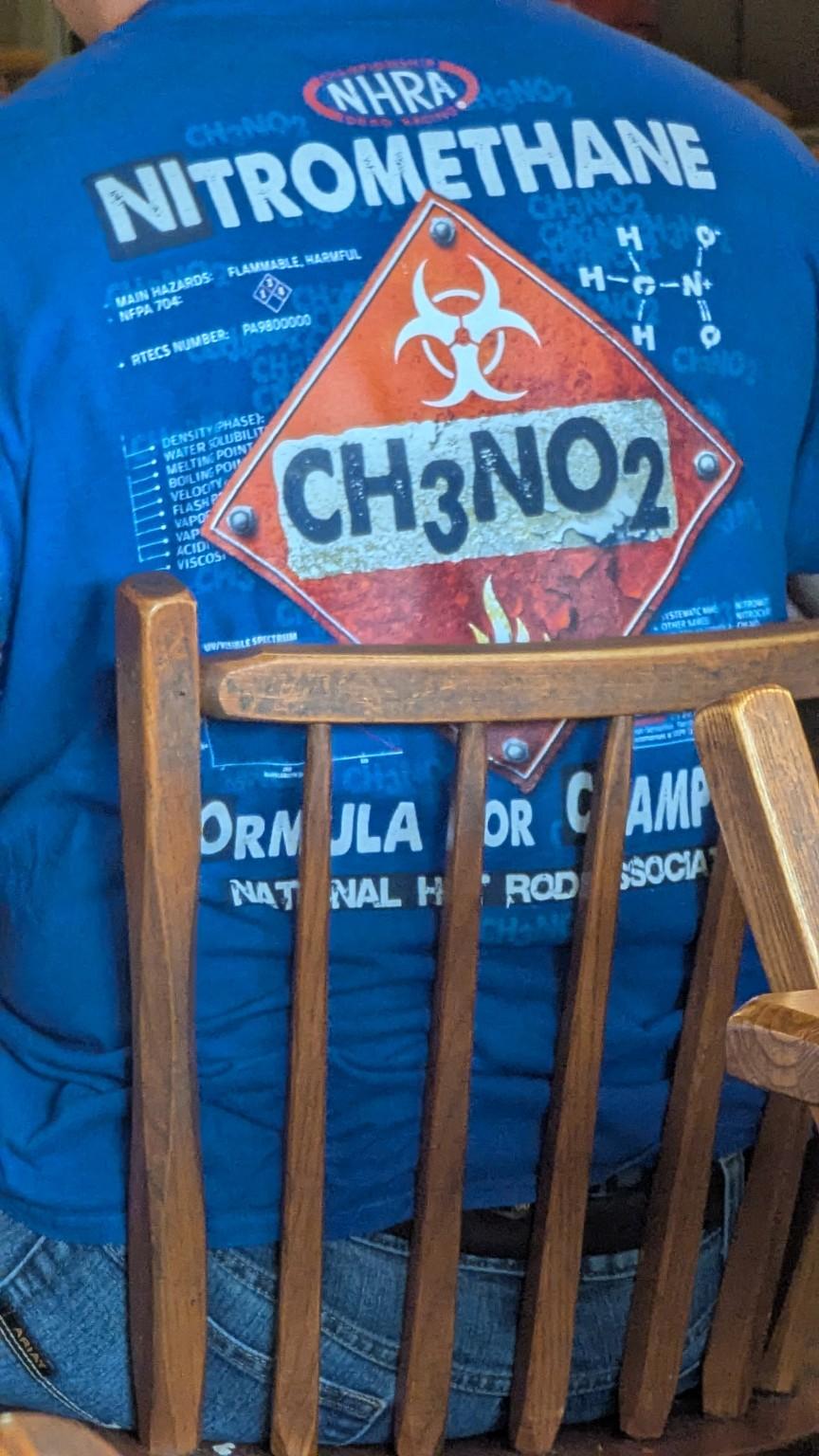 The original phone photograph: the back of a blue NHRA shirt reading "Nitromethane" with a hazard diamond around CH3NO2, partly hidden by a wooden chair back.